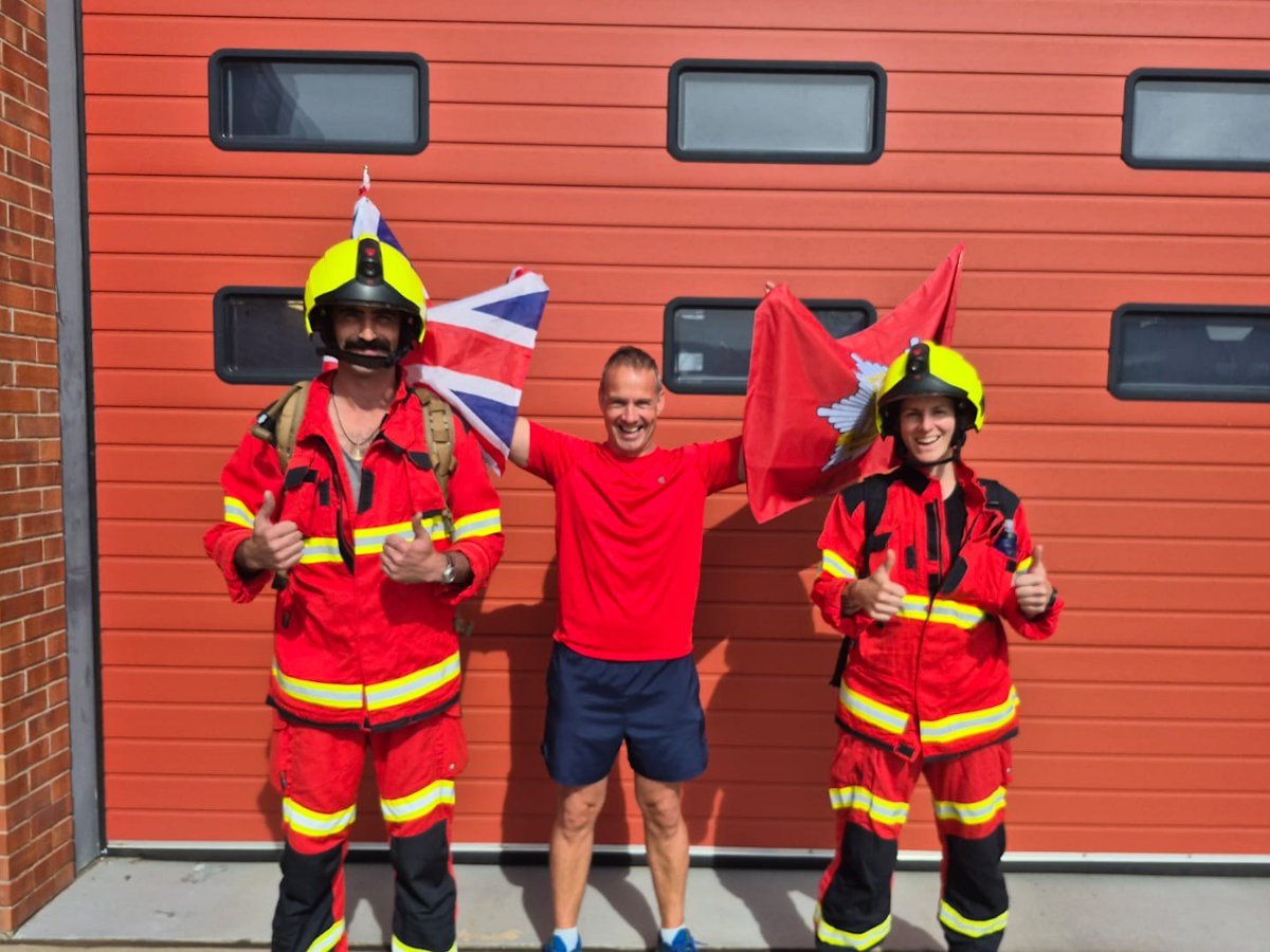 Two firefighters in full protective kit stand either side of a man in running gear holding flags, ahead of their half marathon stretcher challenge